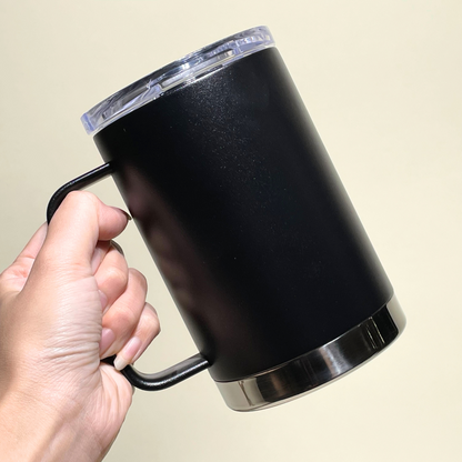 A black stainless steel insulated mug with a clear spill-proof lid, held in hand. This double-wall vacuum-sealed travel mug keeps beverages hot for 6 hours and cold for 24 hours, making it ideal for coffee, tea, or on-the-go drinks. The sturdy ergonomic handle ensures a comfortable grip.
