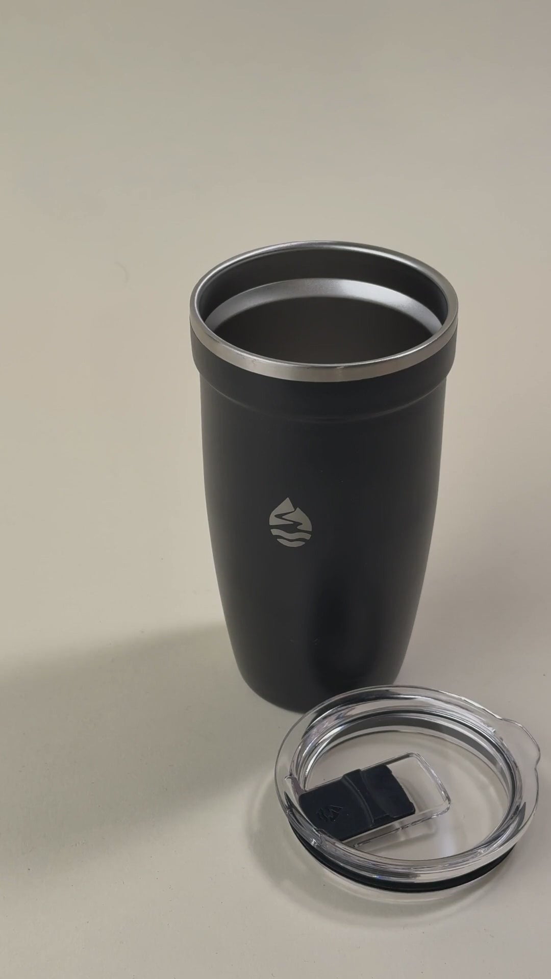 Load video: Black insulated stainless steel tumbler with a clear spill-resistant lid, featuring a sleek design and a minimalist engraved logo.