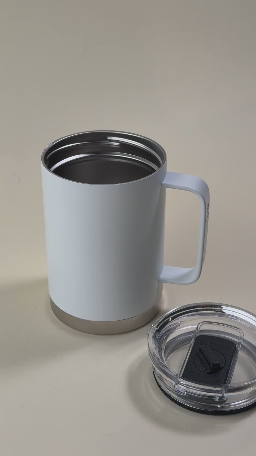 Load video: White insulated stainless steel mug with a sturdy handle, held by a hand, alongside a transparent spill-proof lid.